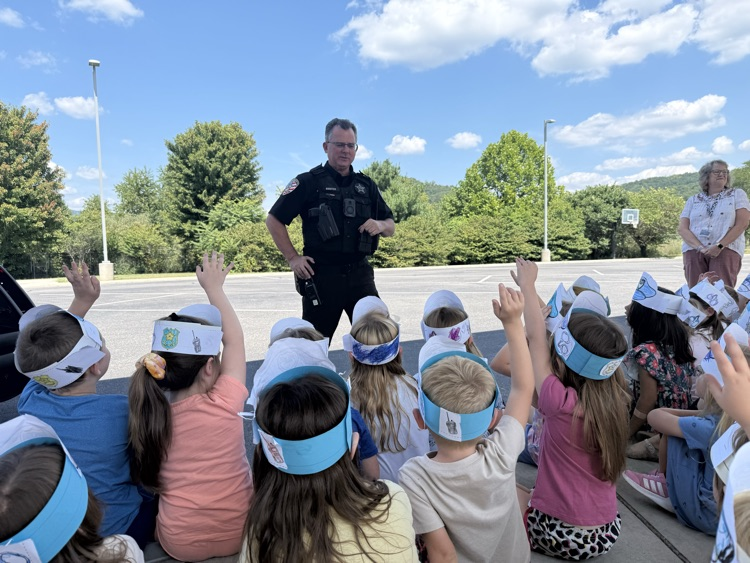 Kindergarten students wearing paper police hats sitting outdoors and raising their hands to ask a school resource officer a question.