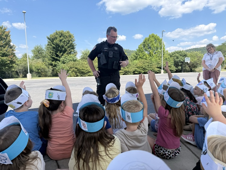 Kindergarten students wearing paper police hats sitting outdoors and raising their hands to ask a school resource officer a question.