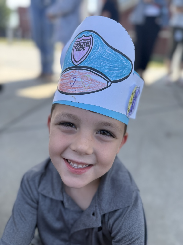 A smiling young boy wearing a paper police hat outdoors.