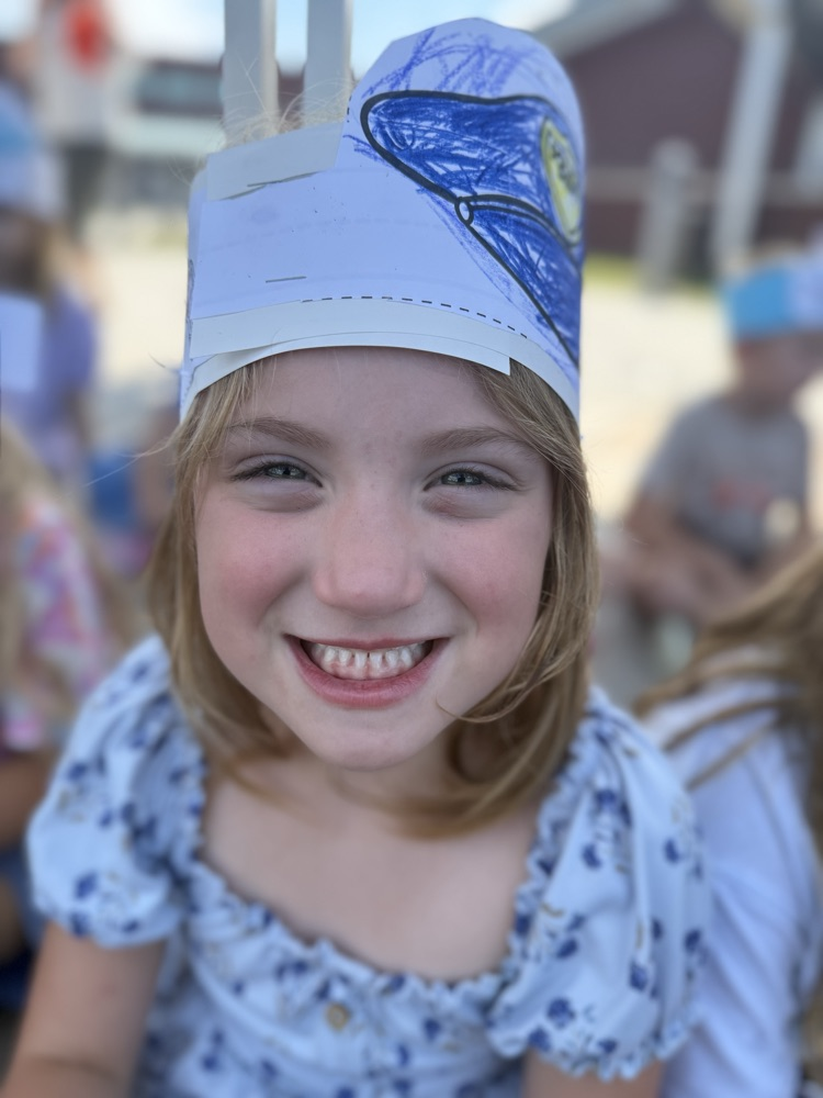A smiling young girl wearing a paper police hat outdoors.