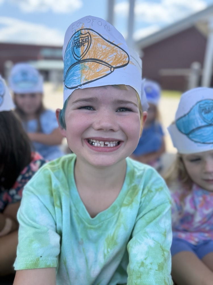A smiling young boy wearing a paper police hat outdoors.
