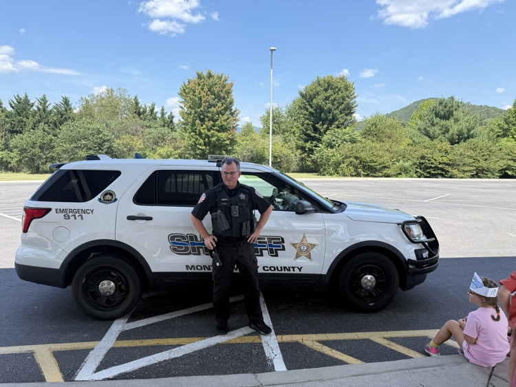 A school resource officer standing outdoors next to a white Sheriff patrol vehicle.