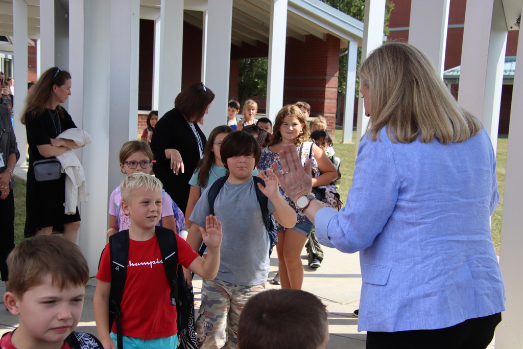 A woman in a light blue blazer exchanges a high-five with a student as children with backpacks walk along a covered school walkway.