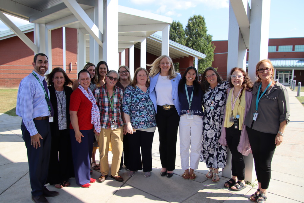 A woman in a light blue blazer stands with a smiling group of school staff beneath a covered walkway outside the school.