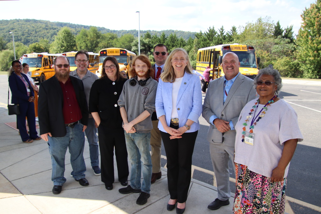 School staff and visitors pose together outdoors in front of several yellow school buses on a sunny day.