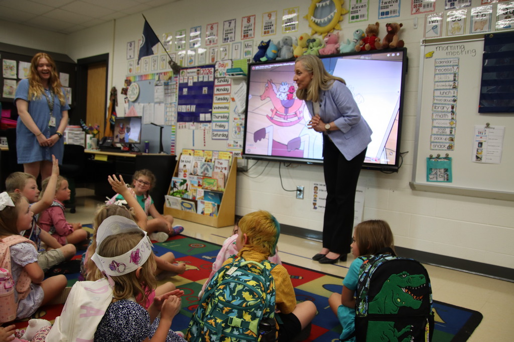 A woman in a light blue blazer speaks with young students seated on a classroom rug as several eagerly raise their hands.