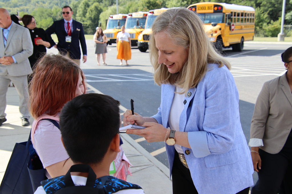 A woman in a light blue blazer signs a notebook for students outside a school, with yellow school buses parked behind them.