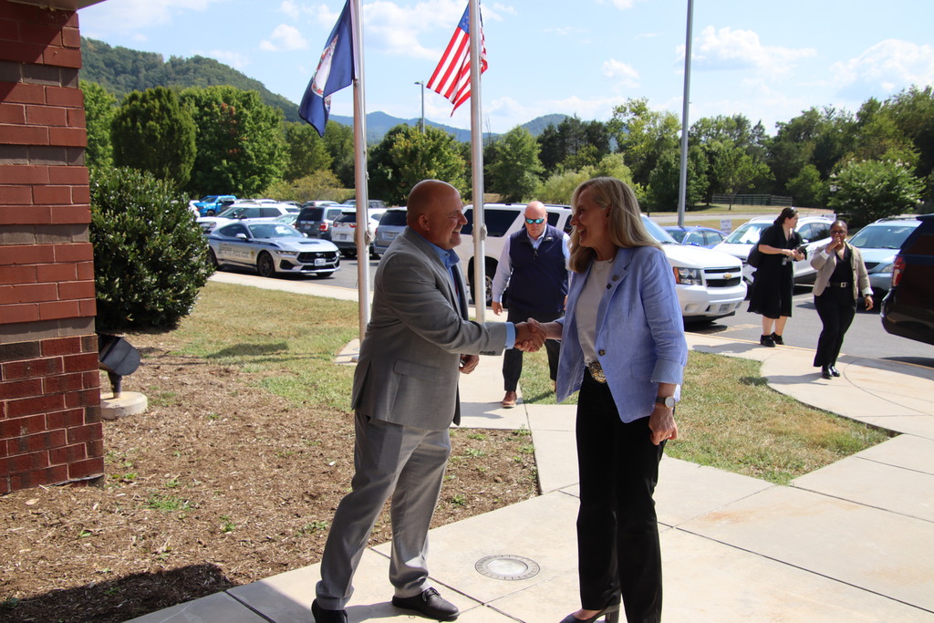 A man in a gray suit and a woman in a light blue blazer smile and shake hands outside a school beneath U.S. and state flags.