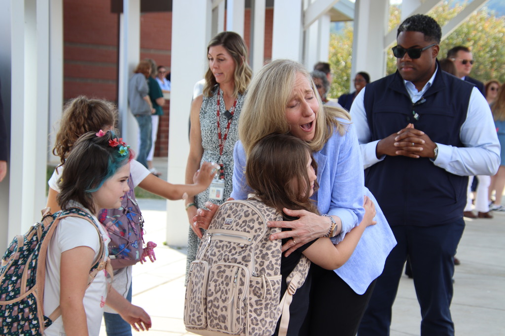 A woman in a light blue blazer warmly hugs a student with a backpack outside a school as staff and students gather nearby.