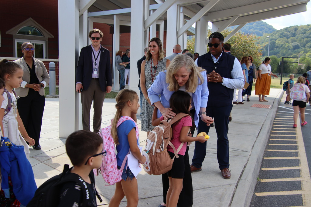 A woman in a light blue blazer bends down to greet and hug a student with a backpack outside the school entrance.