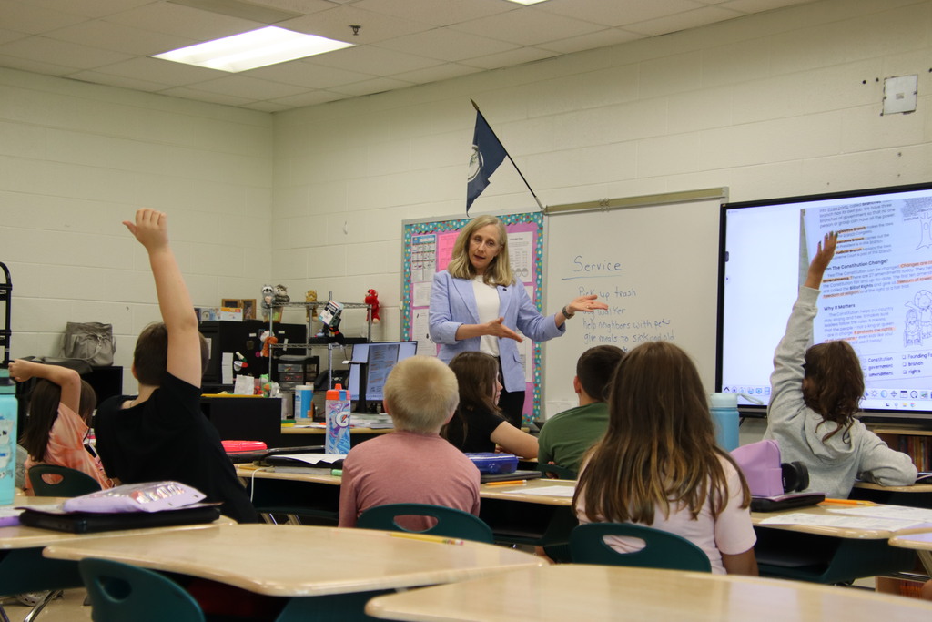 Students raise their hands as a woman in a light blue blazer leads a classroom discussion about service.