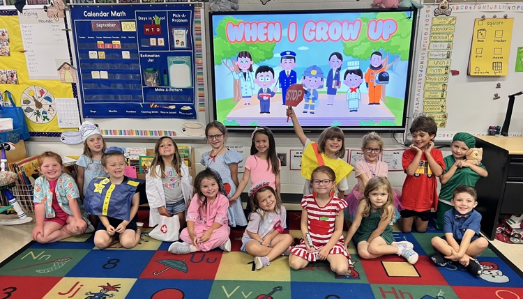 Kindergarten students in career day costumes pose together in two rows on an educational rug inside a classroom. Behind them, a large screen displays a colorful illustration reading "WHEN I GROW UP" alongside various cartoon community helpers.