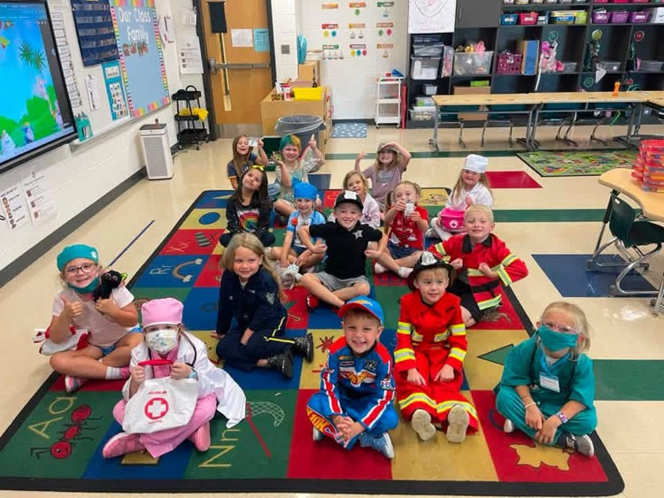 Kindergarten students in career day costumes pose together on an educational rug inside a classroom. 