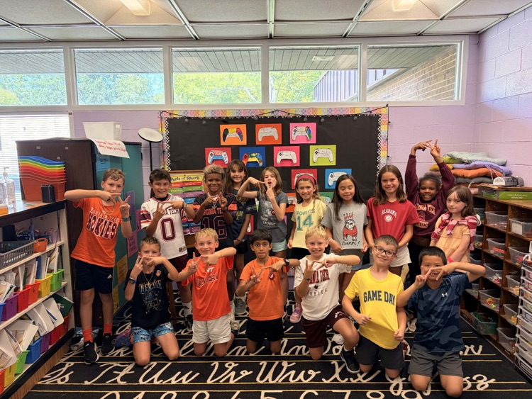 a group of fourth grade students pose in their classroom wearing various college shirts