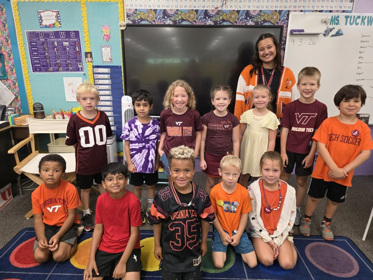 a group of first grade students pose in their classroom wearing various college shirts along with their classroom student teacher 