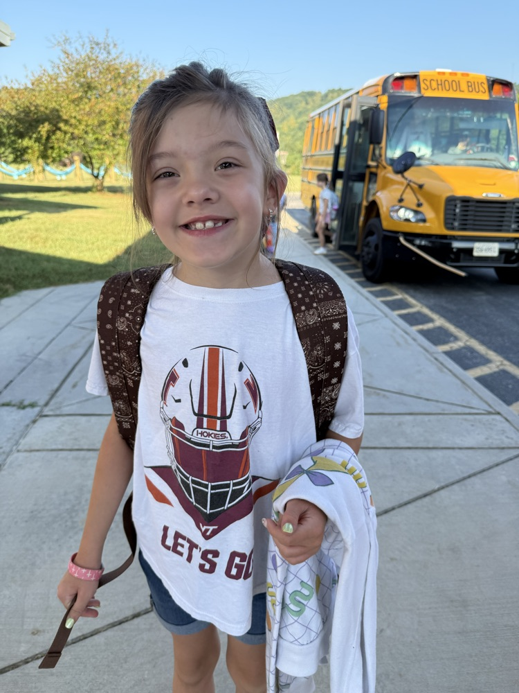 Young girl wearing a white Virginia Tech t-shirt and backpack outdoors near a yellow school bus.
