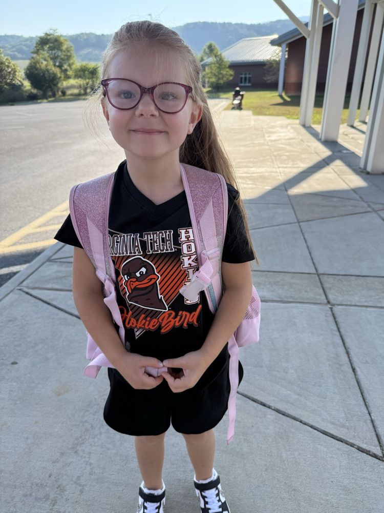 Young girl with glasses and a pink backpack wearing a black Virginia Tech Hokie Bird t-shirt outdoors.