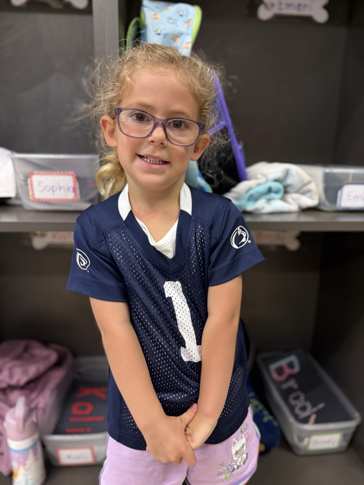 Young girl with glasses wearing a navy blue jersey standing in front of classroom cubbies wearing Penn State jersey.