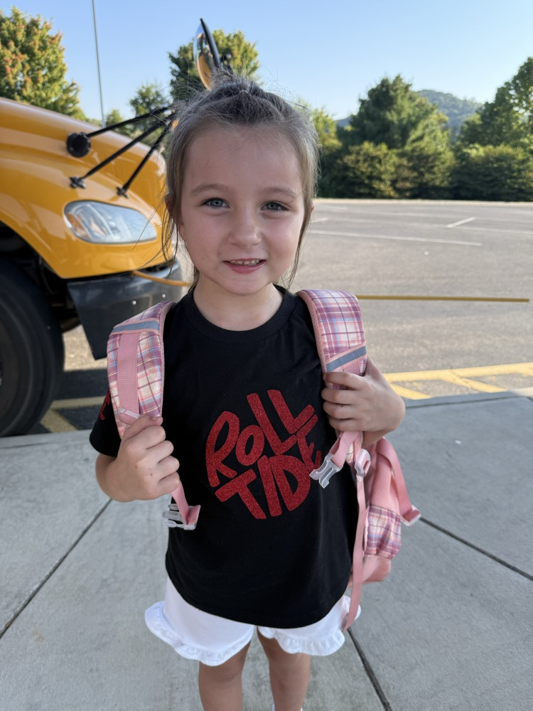 Young girl with a pink backpack wearing a black "Roll Tide" t-shirt outdoors by a school bus.