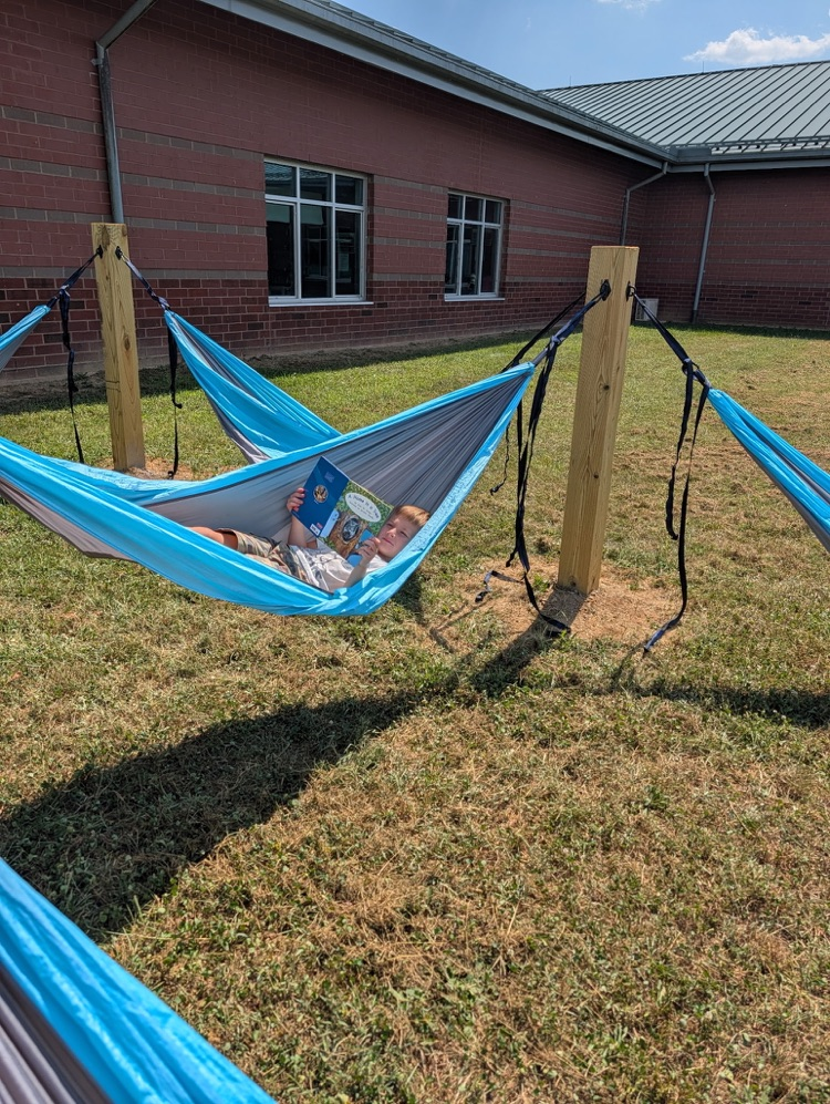 A young boy lying in a hammock while reading a book outdoors.
