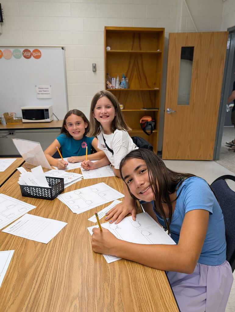 Three students smile while seated at a classroom table completing a literacy activity with pencils and worksheets.