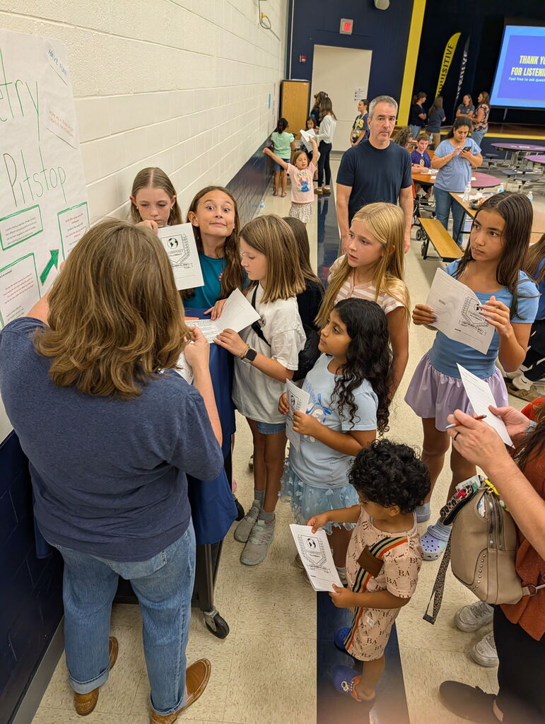 Students and families participate in a Literacy Night activity station in the cafeteria, holding activity sheets and gathering around a staff member for directions.