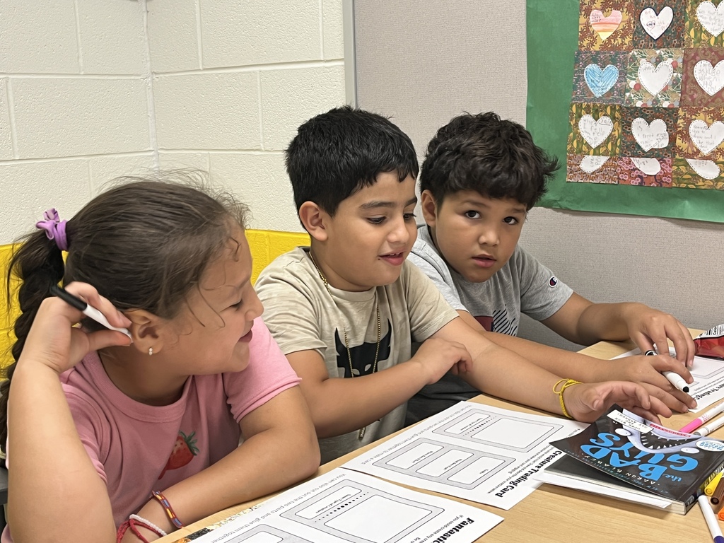 Three elementary students sit together at a table completing literacy activities. They are smiling and talking while working on worksheets with markers.