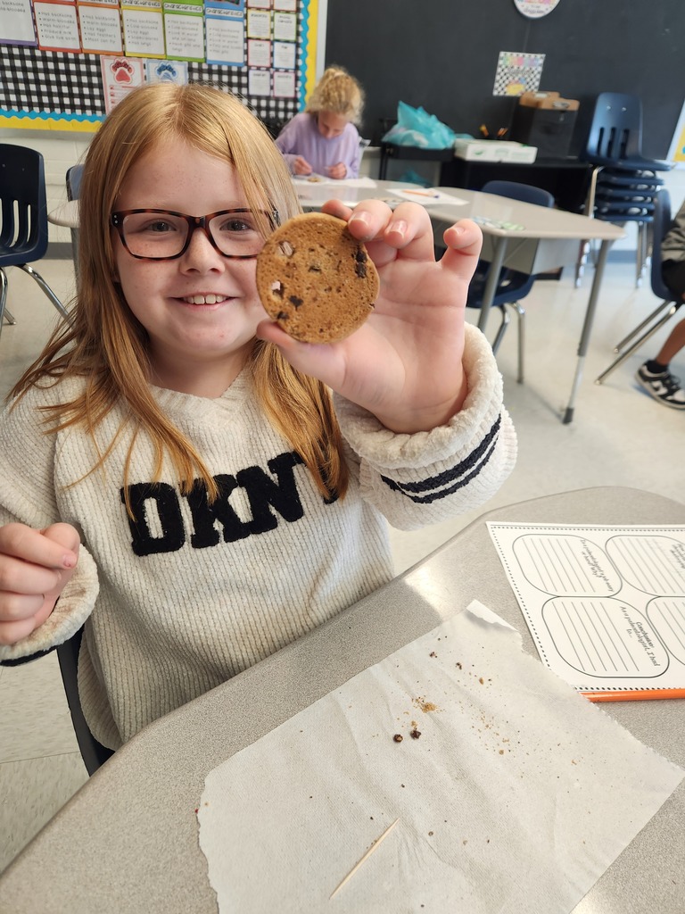 A smiling elementary student sitting at a desk showing her results of simulating recovering fossils with chocolate chip cookies while following the scientific process