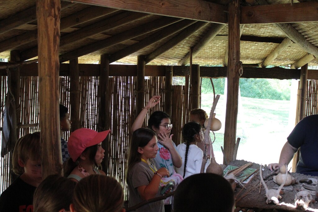 Elementary students in a hut in a native american village museum asking questions to their tour guide.