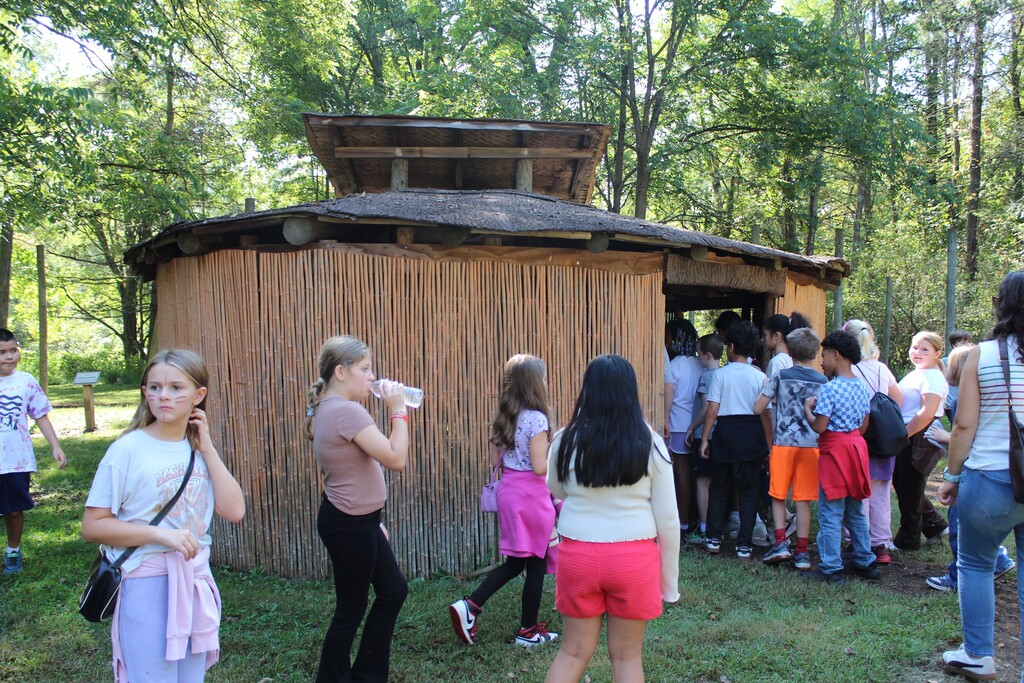 A group of elementary students on a field trip to a native american museun entering a hut.
