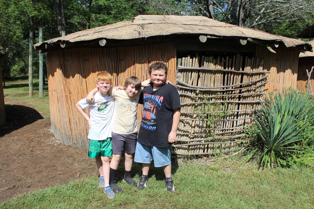 Three students posing outside a hut at a native american indian village and museum.
