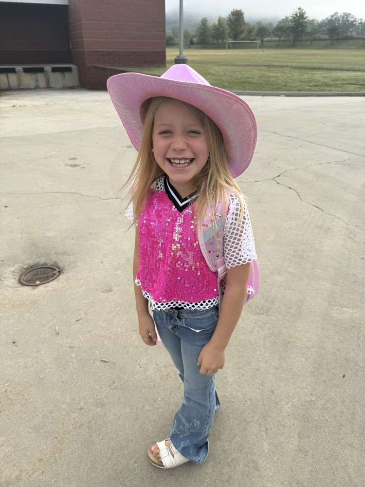 A smiling girl wearing a bright pink cowboy hat, a pink sequined top over a mesh shirt, and flare jeans.