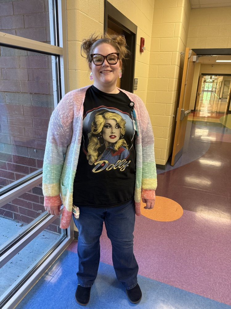 A smiling woman with her hair in twin buns wearing a pastel rainbow cardigan and a black graphic tee featuring Dolly Parton.
