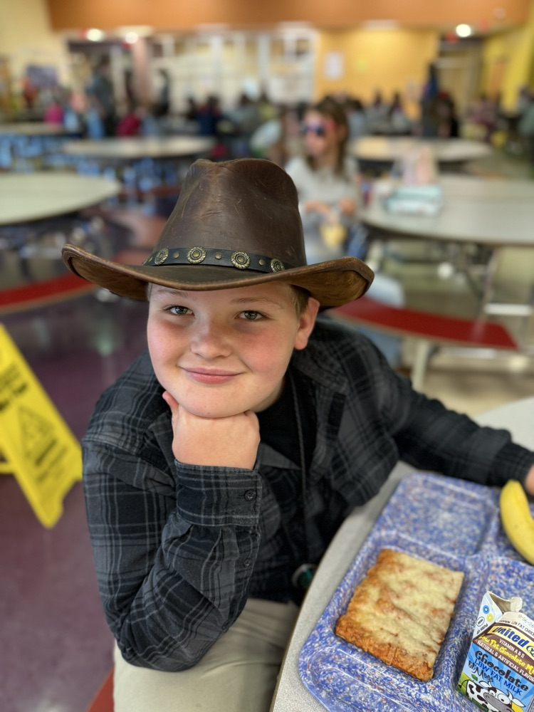 A boy wearing a leather cowboy hat and a black plaid shirt leaning his chin on his hand at a lunch table.