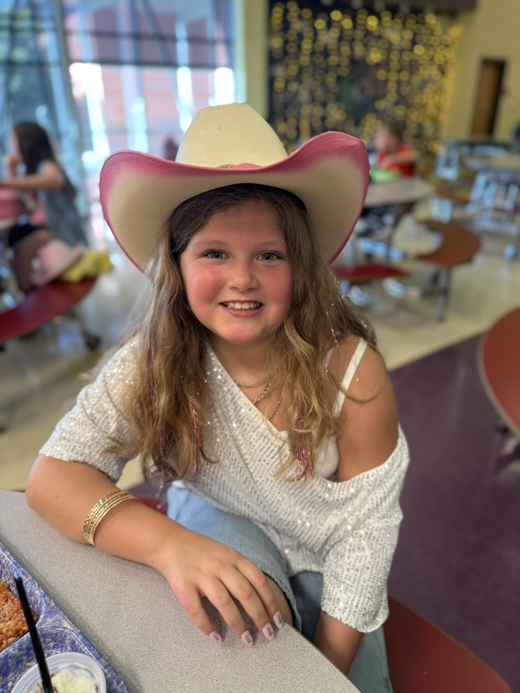 A smiling girl sitting at a table wearing a pink-brimmed cowboy hat and a sparkly white off-the-shoulder shirt.