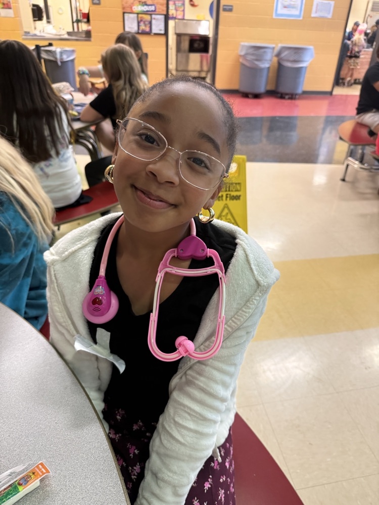 A young girl with glasses smiling warmly while wearing a pink toy stethoscope around her neck.