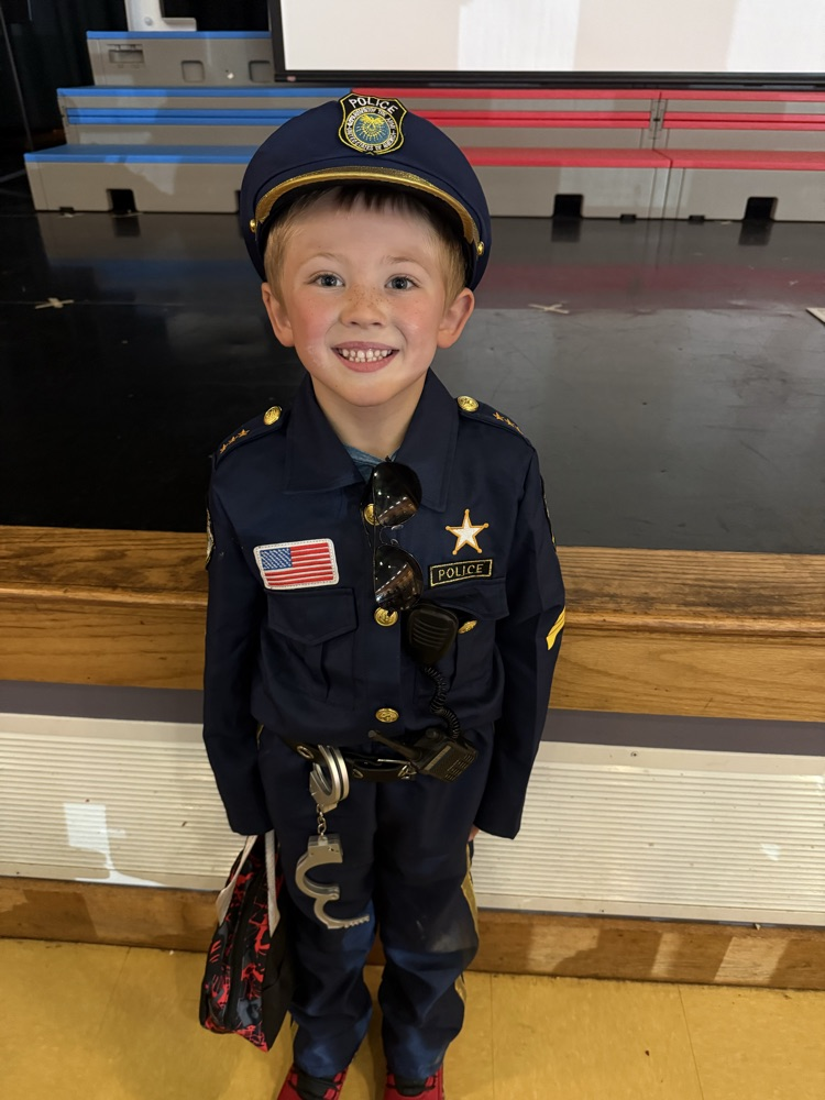 A young boy smiling in a detailed police officer costume complete with a hat, badge, and handcuffs.