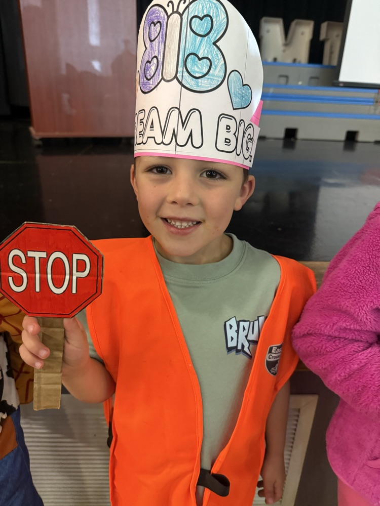 A young boy smiling while holding a small toy stop sign, wearing a high-visibility orange vest and a paper hat decorated with butterflies and the words "Dream Big".