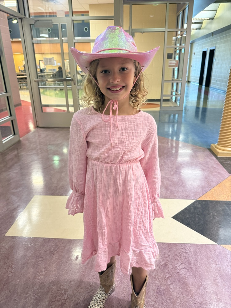A smiling girl wearing a light pink long-sleeved dress, a shimmering pink cowboy hat, and patterned cowboy boots in a hallway.