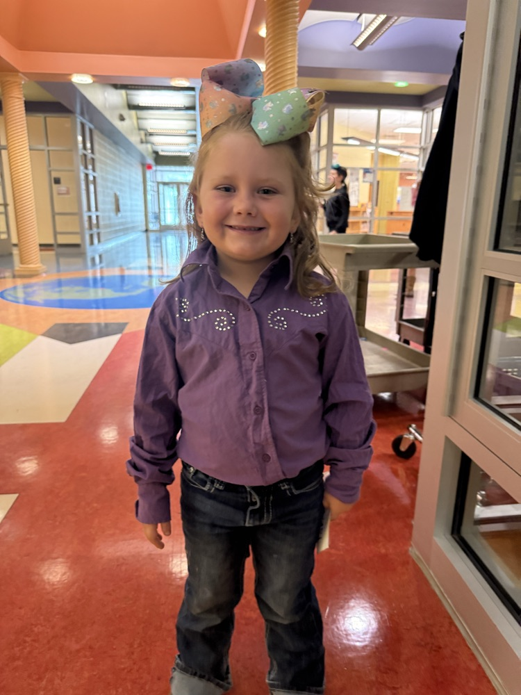 A smiling young girl standing in a school hallway wearing a purple western-style shirt with rhinestone accents, dark jeans, and a large patterned hair bow.