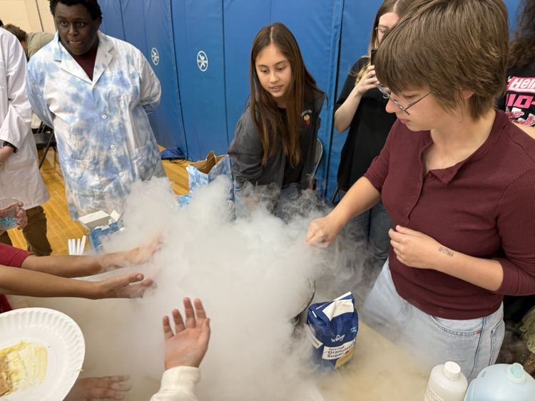 Students watching a science experiment involving thick fog from dry ice spreading across a table.