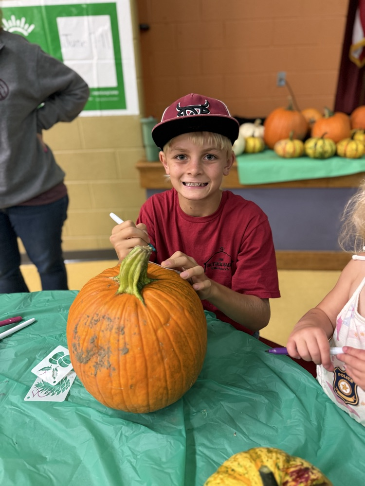 A smiling young boy in a red shirt writing on a large orange pumpkin with a marker.