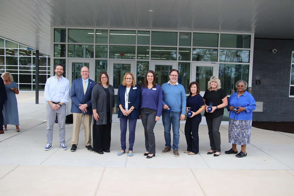 Nine school and community representatives pose outside the school's glass entrance, several holding commemorative scissors.