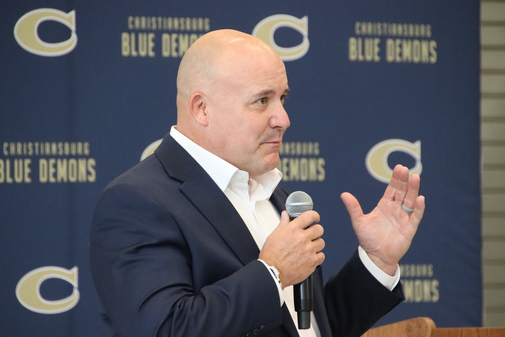 A man in a navy suit speaks into a microphone before a Christiansburg Blue Demons backdrop.