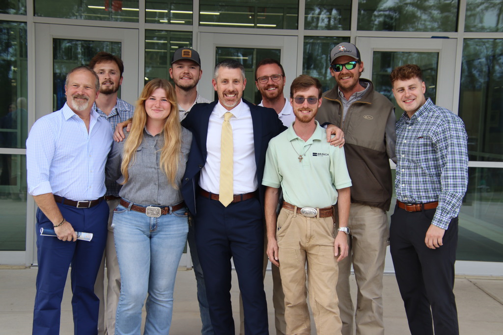 Eight smiling attendees pose together outside the school's glass entrance, including a man in a navy suit and yellow tie.