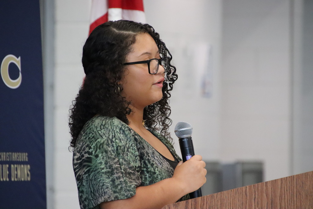 A young woman with curly hair and glasses speaks into a microphone at a podium during the school ceremony.