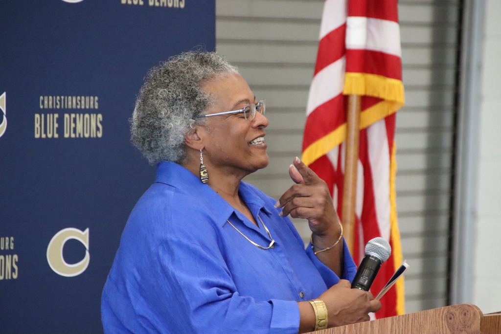 A woman in a bright blue blouse speaks at a podium, with an American flag and Blue Demons banner behind her.