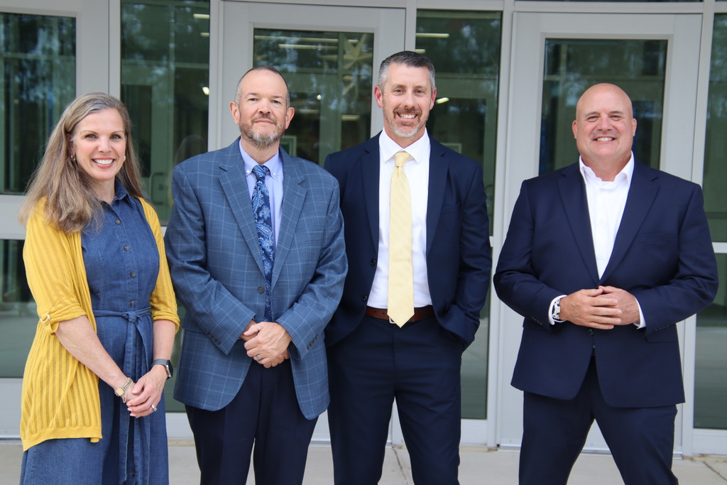 Four smiling school and community representatives pose in professional attire outside the school's glass doors.