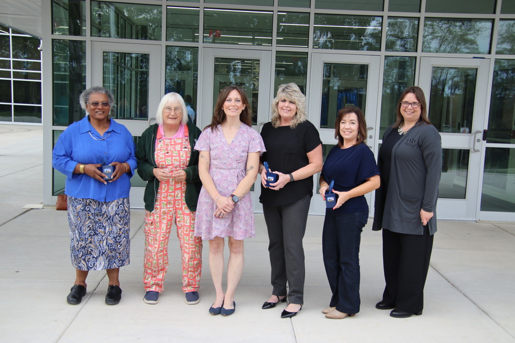 Six women pose outside the school entrance, with three holding commemorative blue scissors in presentation boxes.