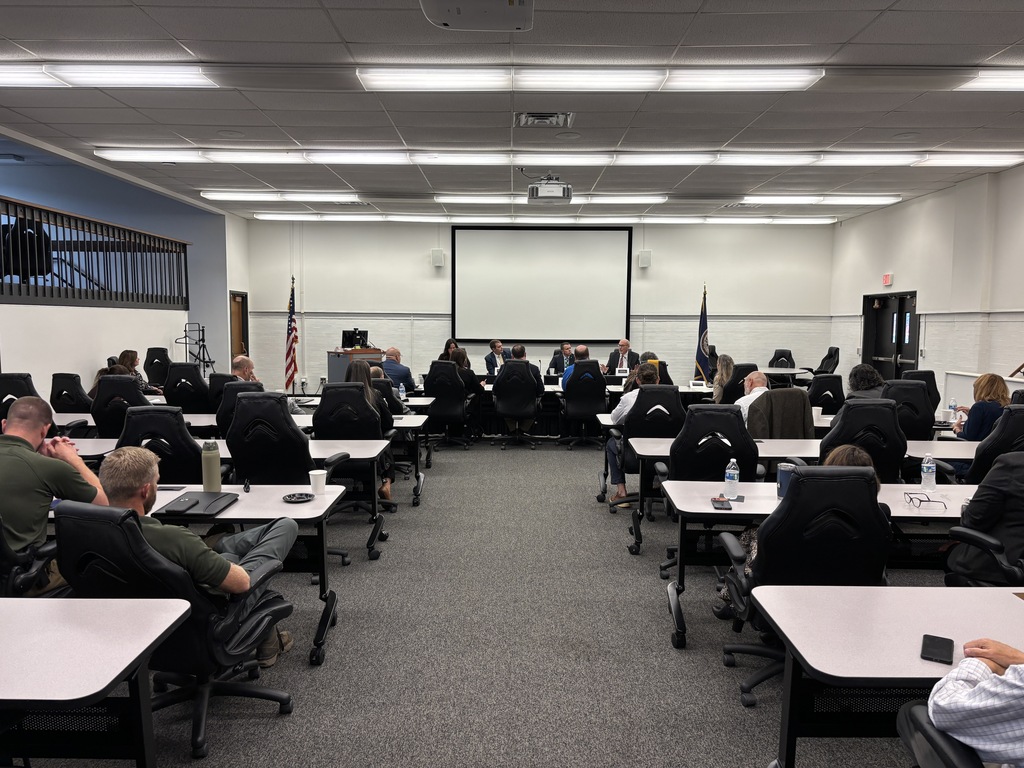 Wide view of a large meeting room filled with attendees seated at rows of tables, facing a panel of speakers and a projection screen at the front during a legislative meeting.